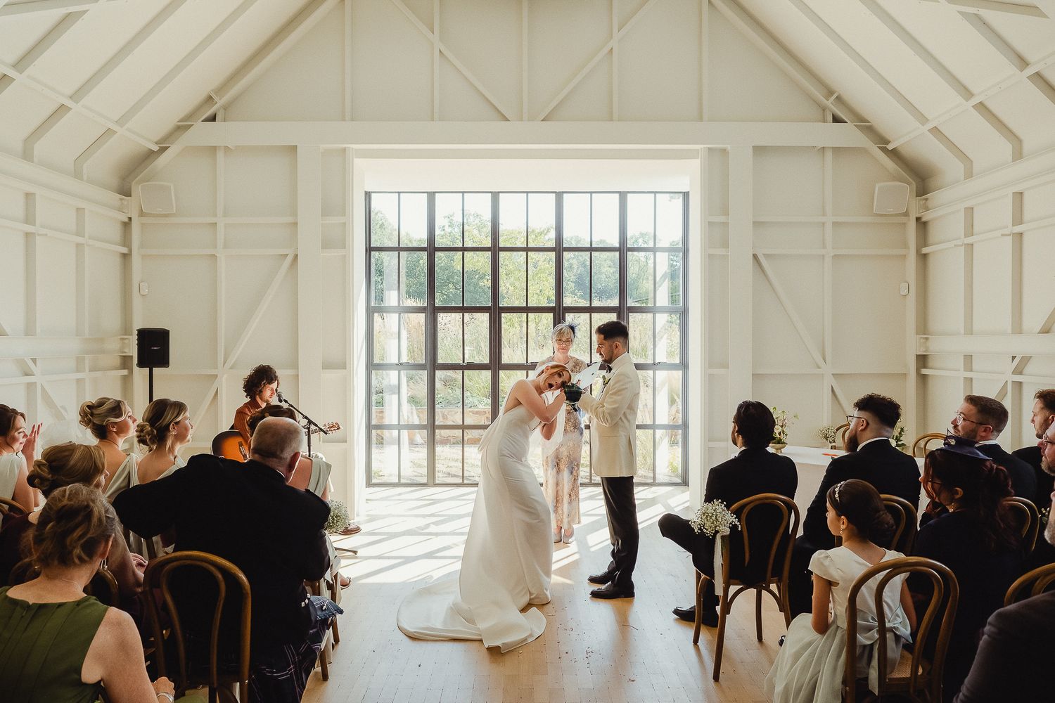 A bright white wedding ceremony venue with large windows and guests seated in rows during a celebration.