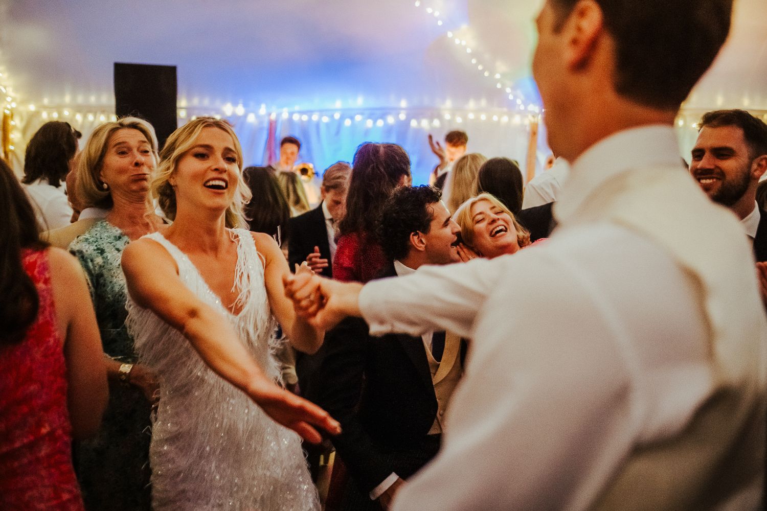 Wedding guests dance and celebrate under twinkling lights at evening reception.