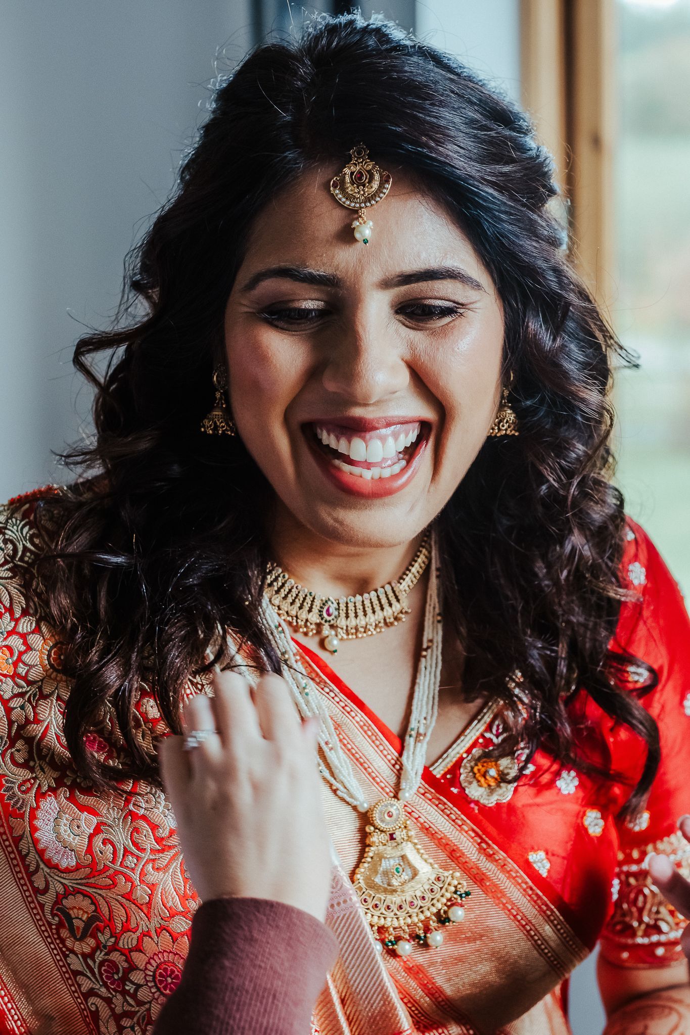 A woman in traditional Indian bridal attire with gold jewelry smiles radiantly during wedding preparations.