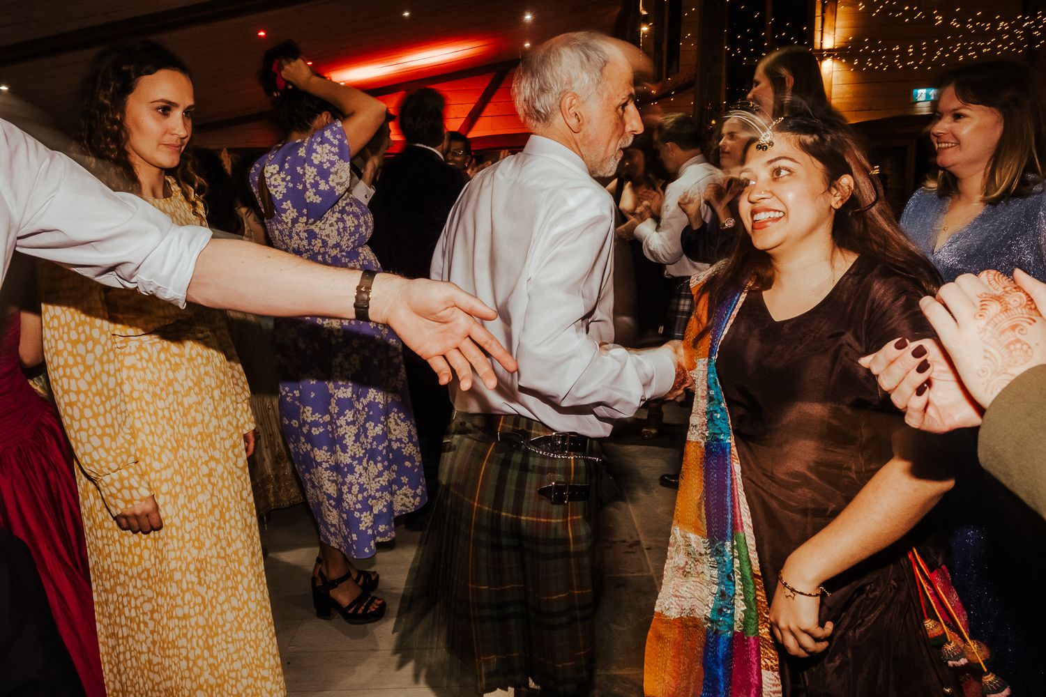 Wedding guests dancing with a person in traditional Scottish kilt at a reception.