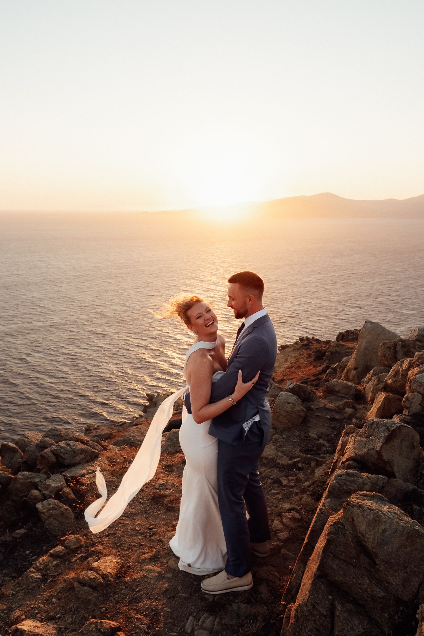 Bride and groom embrace on rocky cliff at golden sunset over ocean, wedding veil flowing in wind.