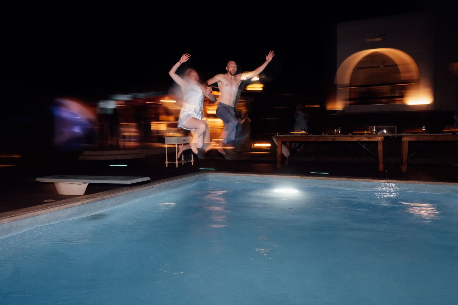 Two people jumping into an illuminated outdoor swimming pool at night.