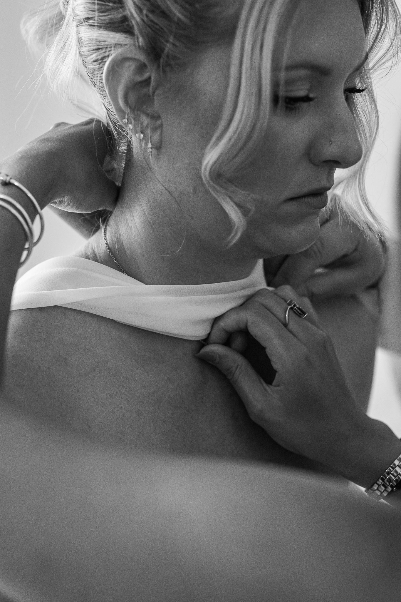 Black and white photo of a bride having her dress fastened, wearing elegant earrings and a delicate necklace.