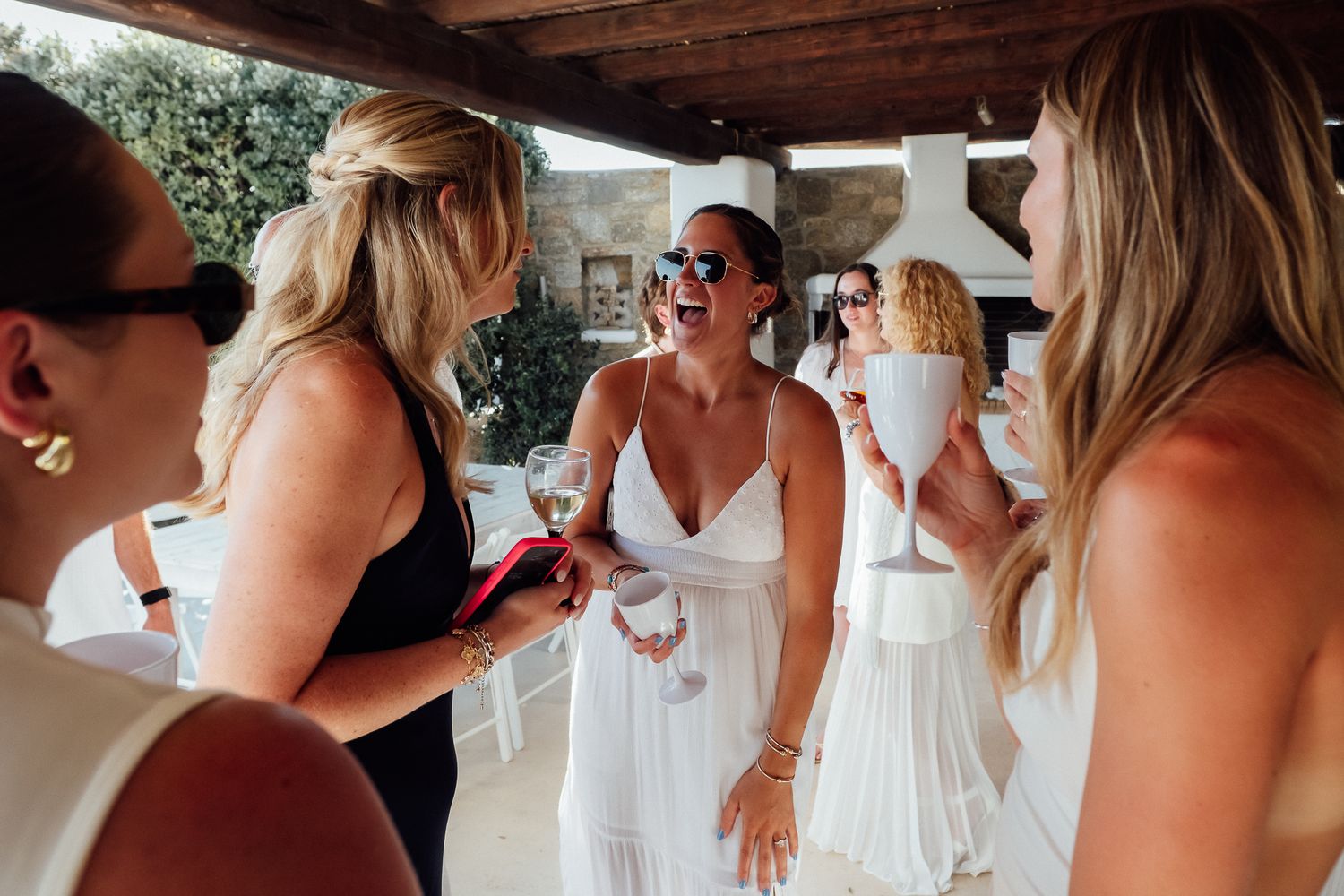 Women in white dresses socializing outdoors at a summer party, holding drinks and smiling.