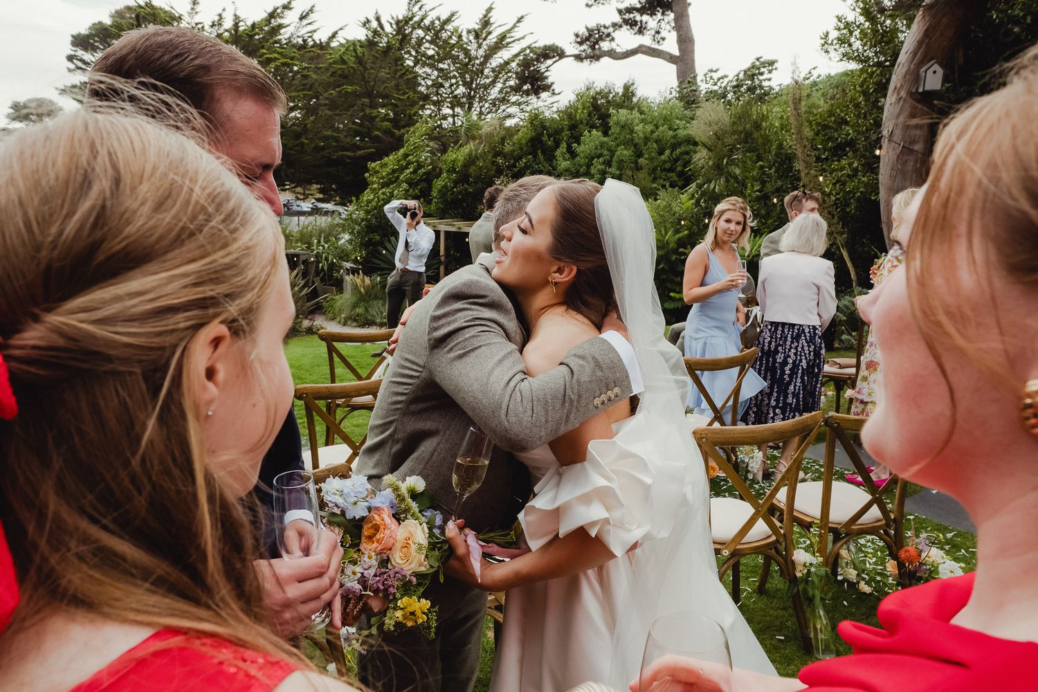 A group of wedding guests gather for an outdoor celebration in a garden setting.