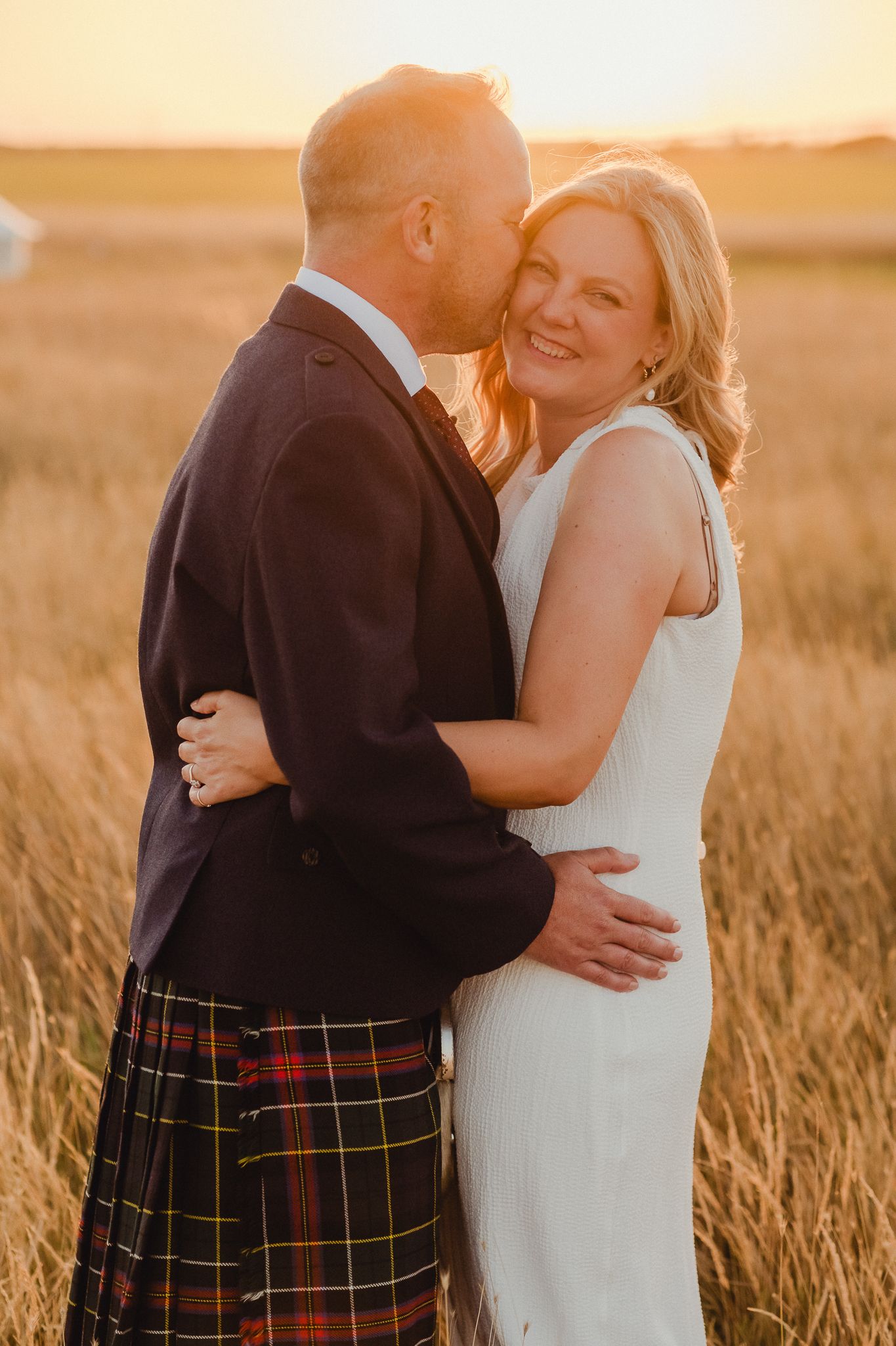 Romantic sunset wedding photos showing couple embracing in golden wheat field with groom wearing traditional Scottish kilt.