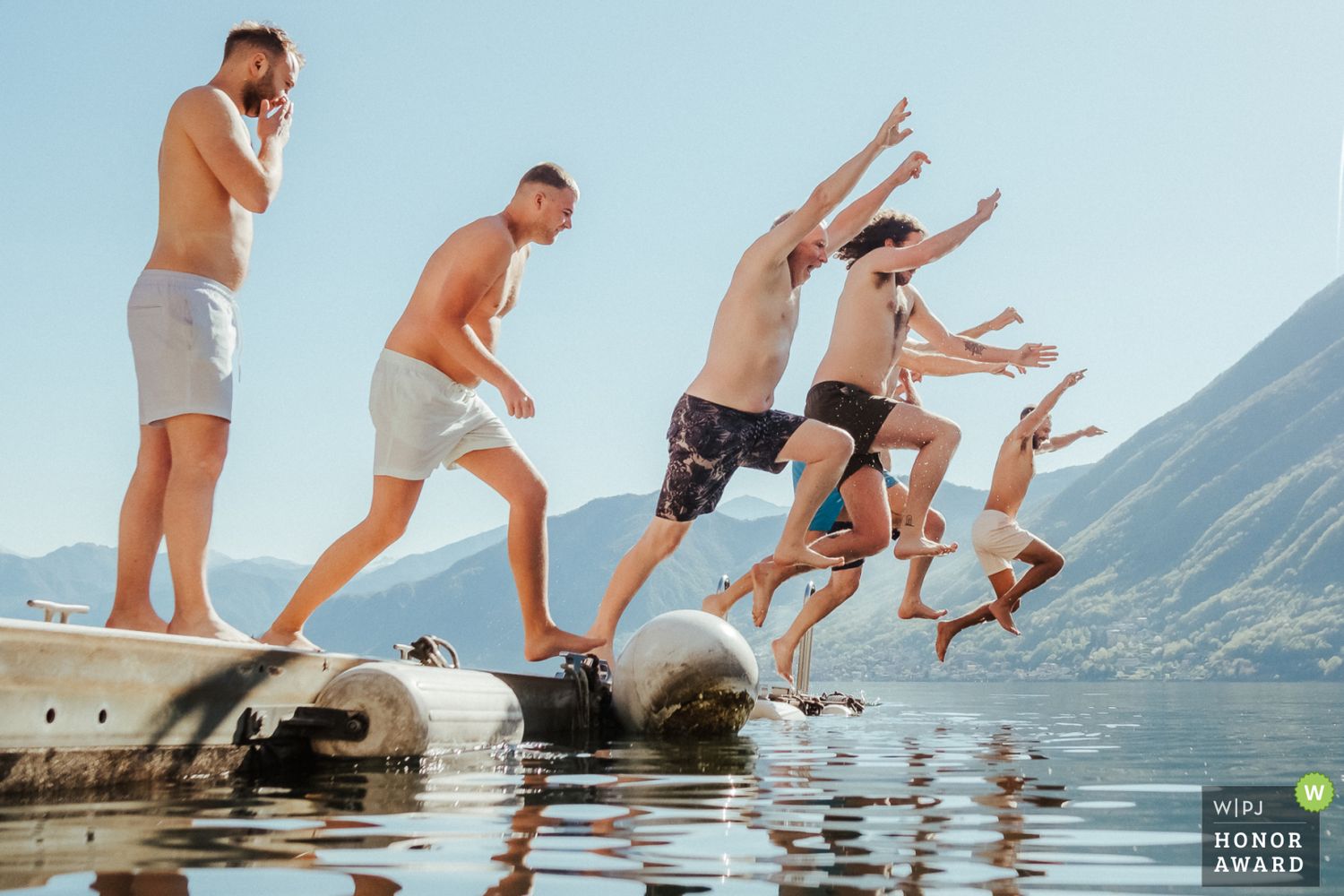A group of friends jumping in sequence off rocks into a scenic mountain lake on a sunny day.