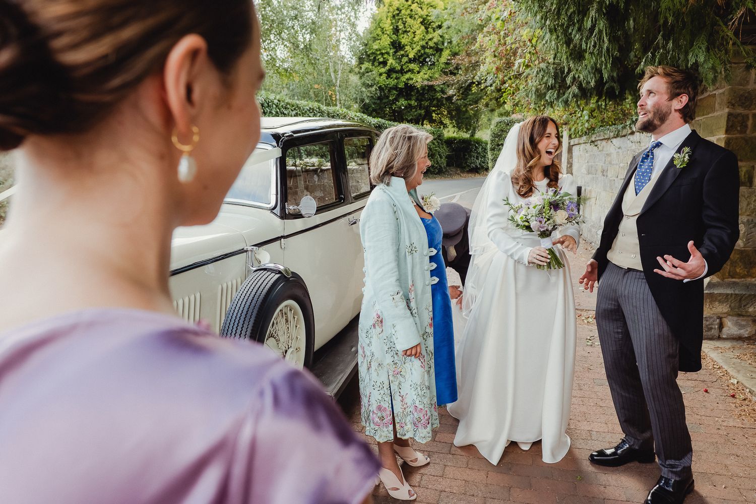 Wedding party gathered outside a vintage cream-colored car on a sunny day.