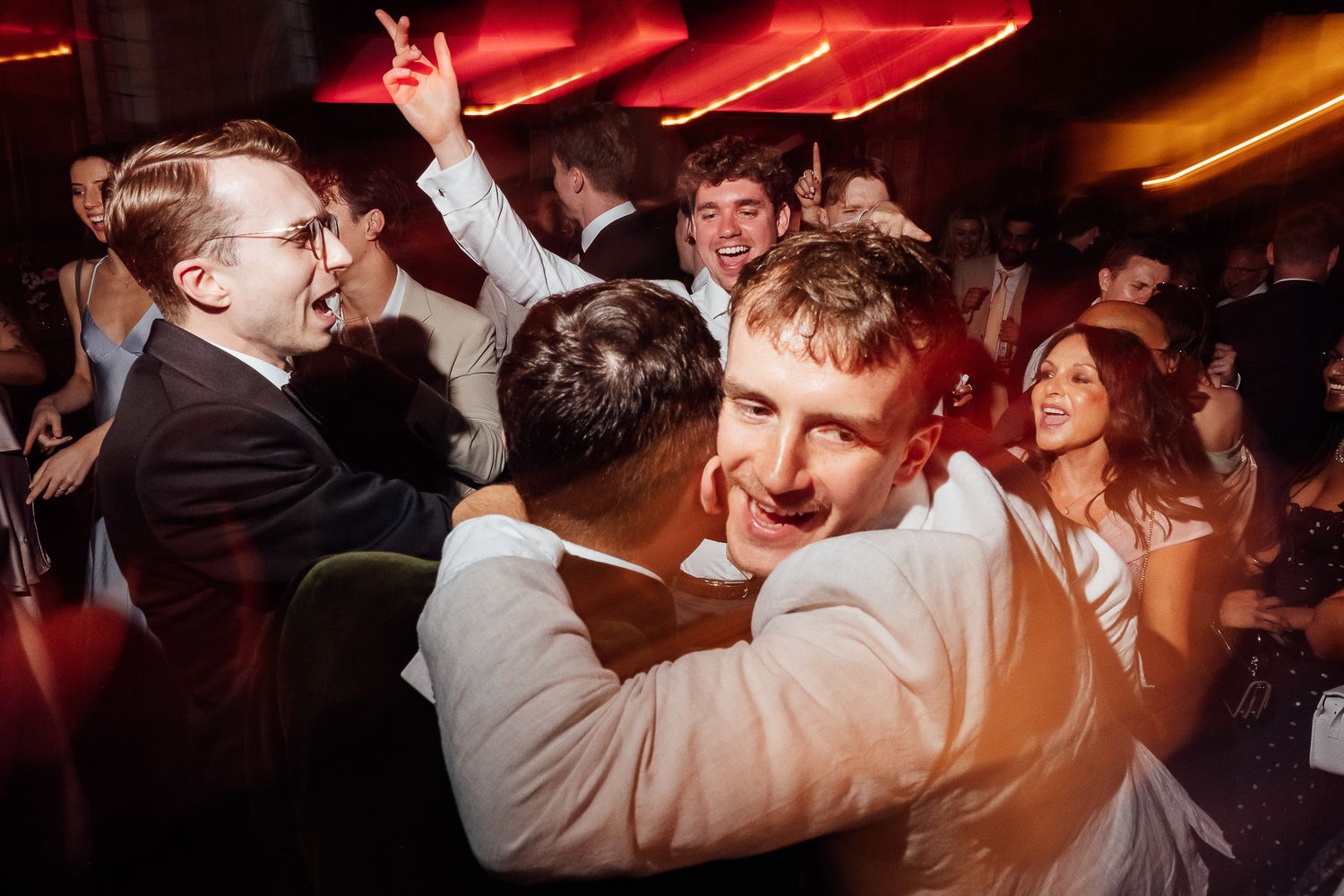 Guests dancing and embracing joyfully at a lively wedding reception party with warm red ambient lighting.