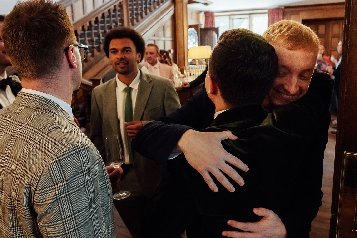 Men in formal attire socializing at an indoor gathering, with two sharing a warm embrace in the foreground.