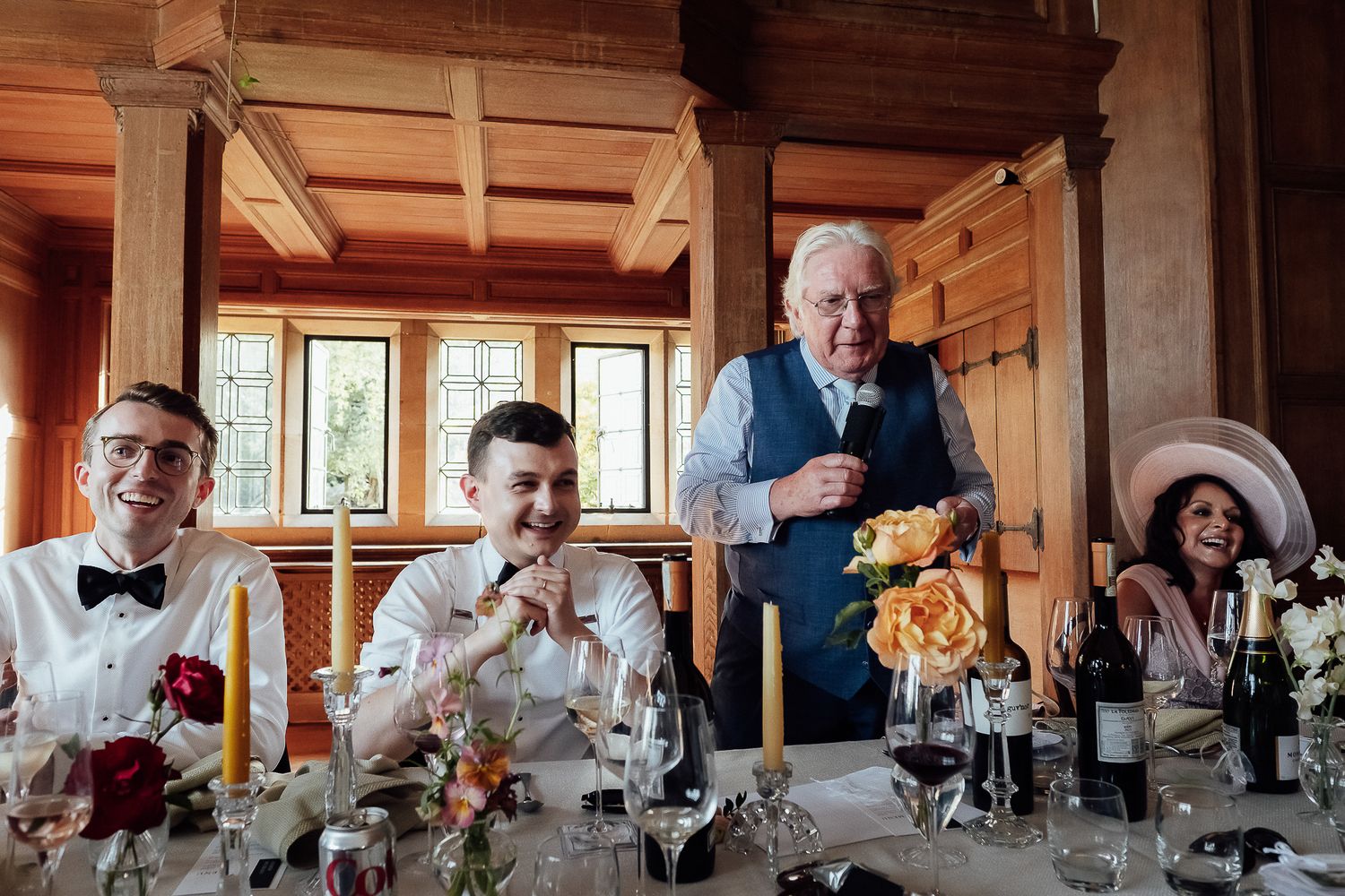 Guests laughing at wedding reception head table with floral centerpieces, candles, and wine glasses in elegant wood-paneled venue.