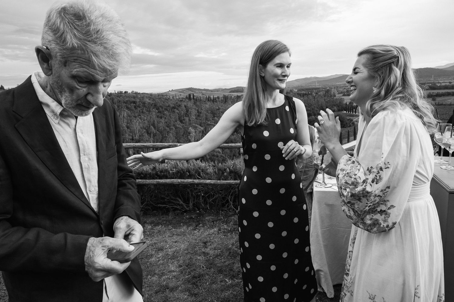 Black and white photo of an older man and two women laughing at an outdoor wedding reception with rolling hills in the background.