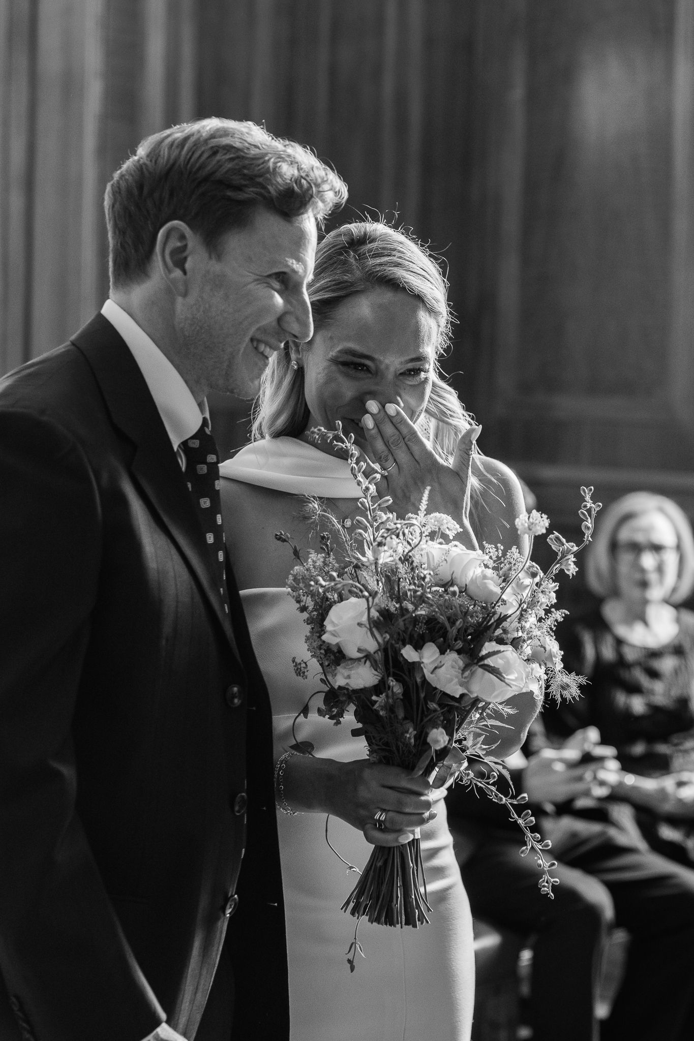 Black and white photo of a smiling groom beside an emotional bride holding a wildflower bouquet during a wedding ceremony.