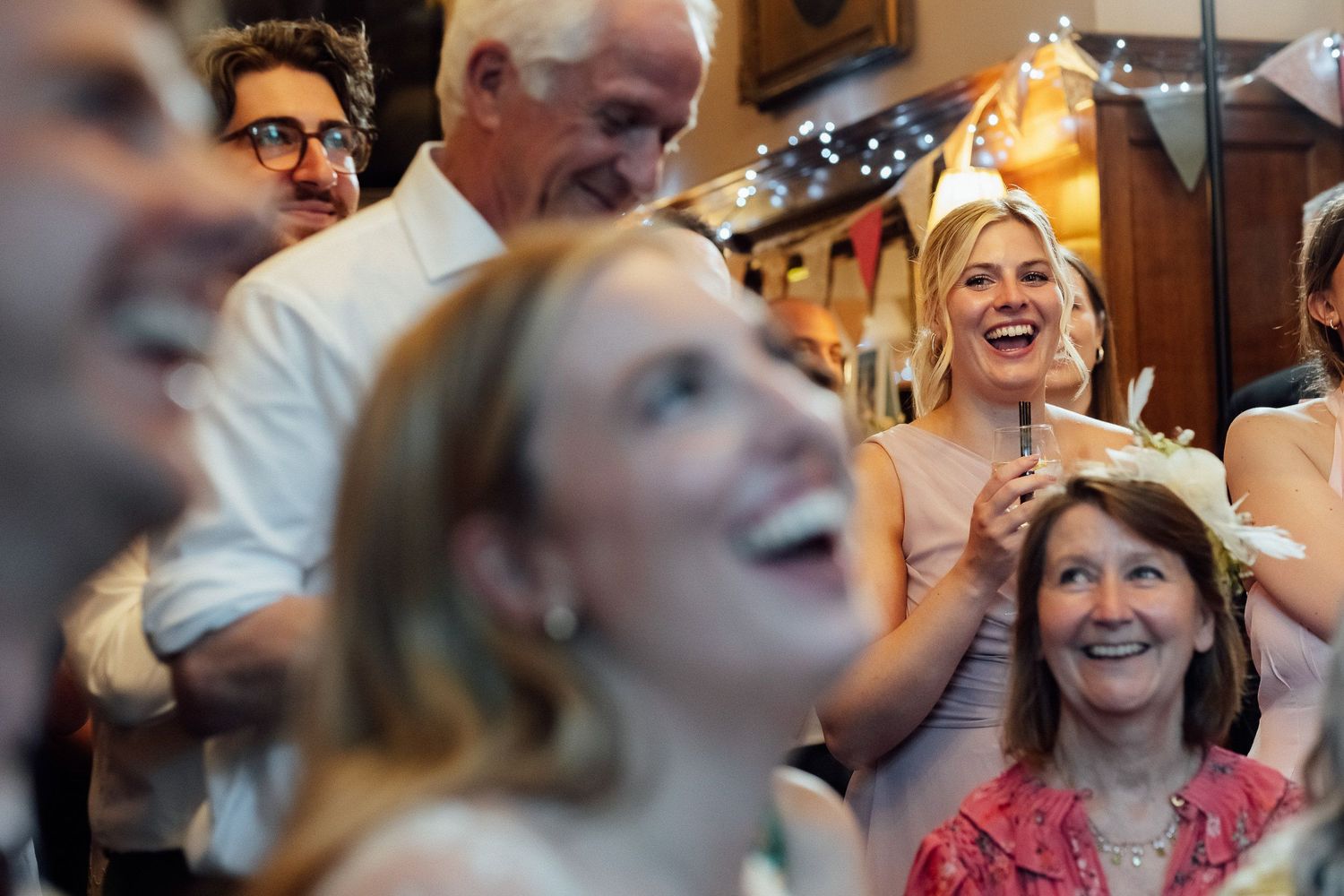 A joyful crowd of guests laughs and smiles together at an indoor celebration under warm string lights, with a woman wearing a floral crown.