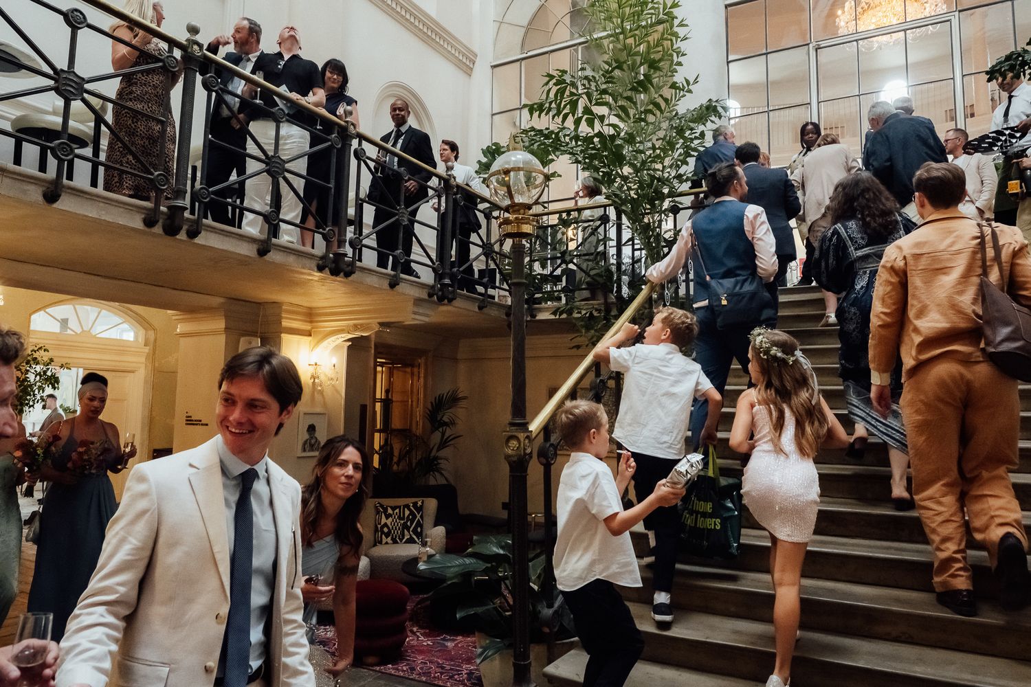 Wedding guests in formal attire climb a grand staircase with children while a smiling man in a light suit stands in the foreground.