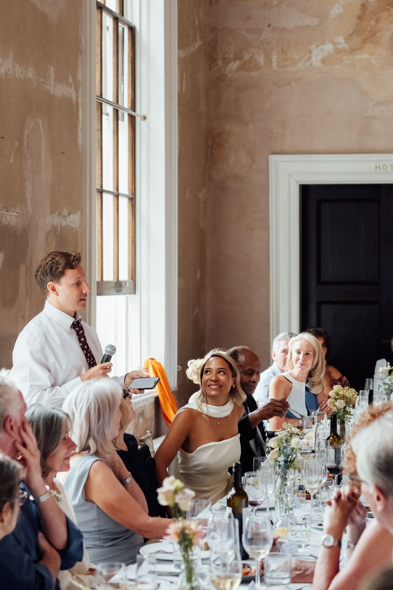 Man in white shirt gives a toast with a microphone as a smiling bride in a white dress and wedding guests sit at a flower-decorated table.