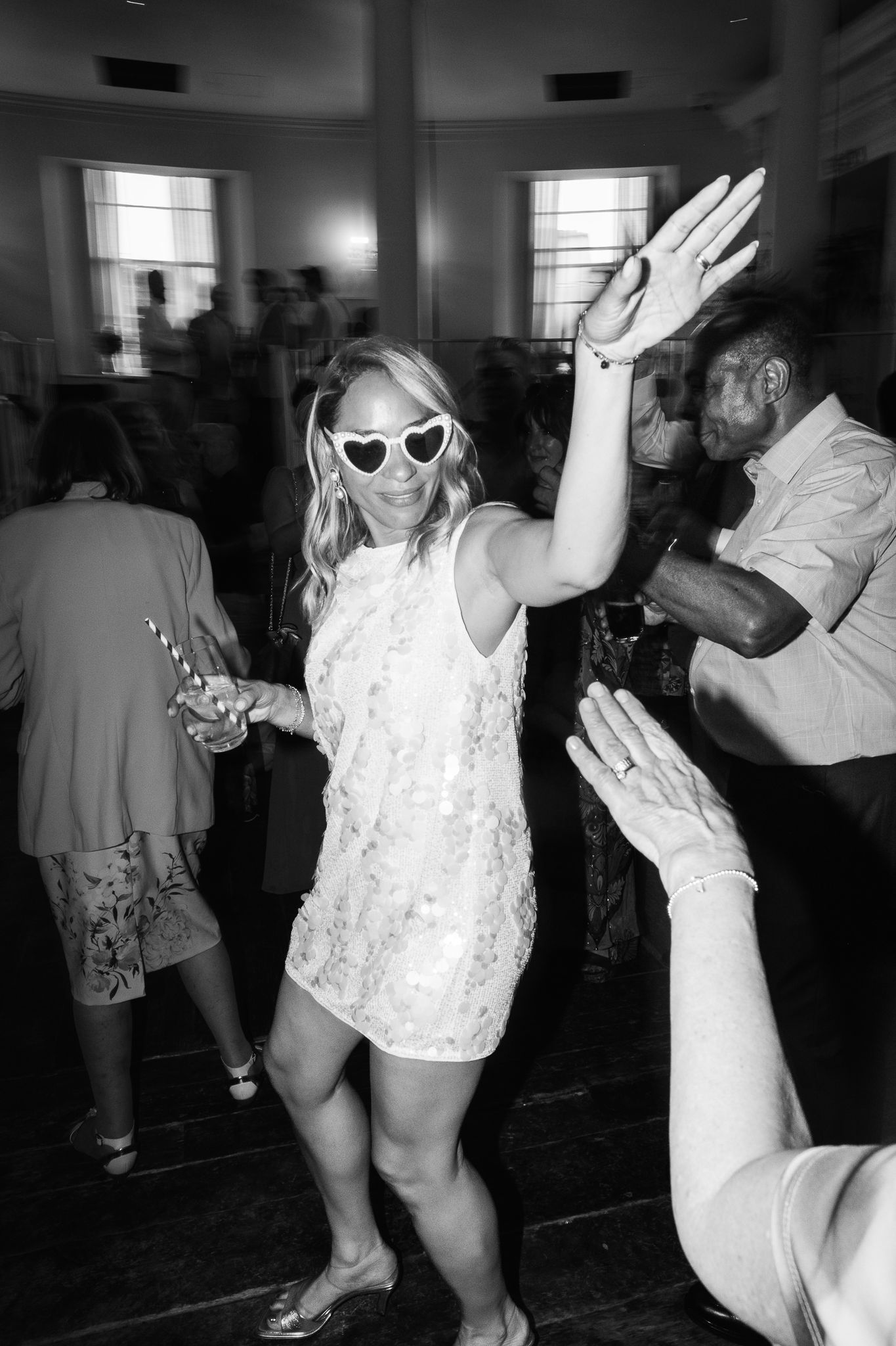 Black and white photo of a woman in a sequined mini dress and heart-shaped sunglasses dancing with her hand raised at a lively party.
