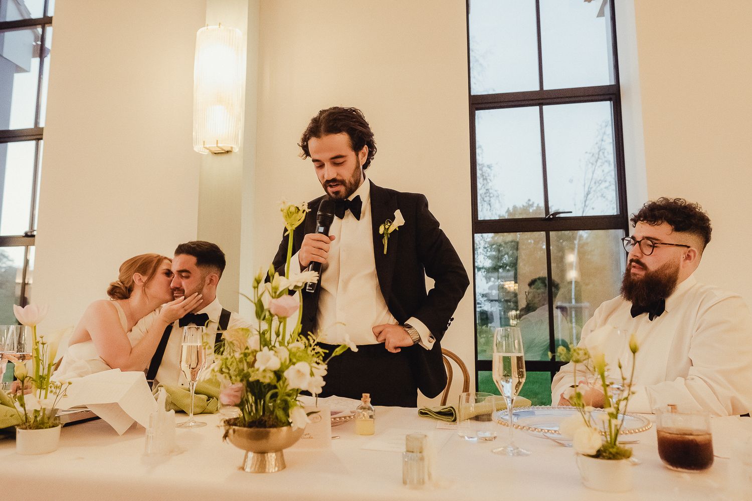 Wedding guests seated at a reception table listen to a speaker giving a toast in an elegant venue with tall windows.