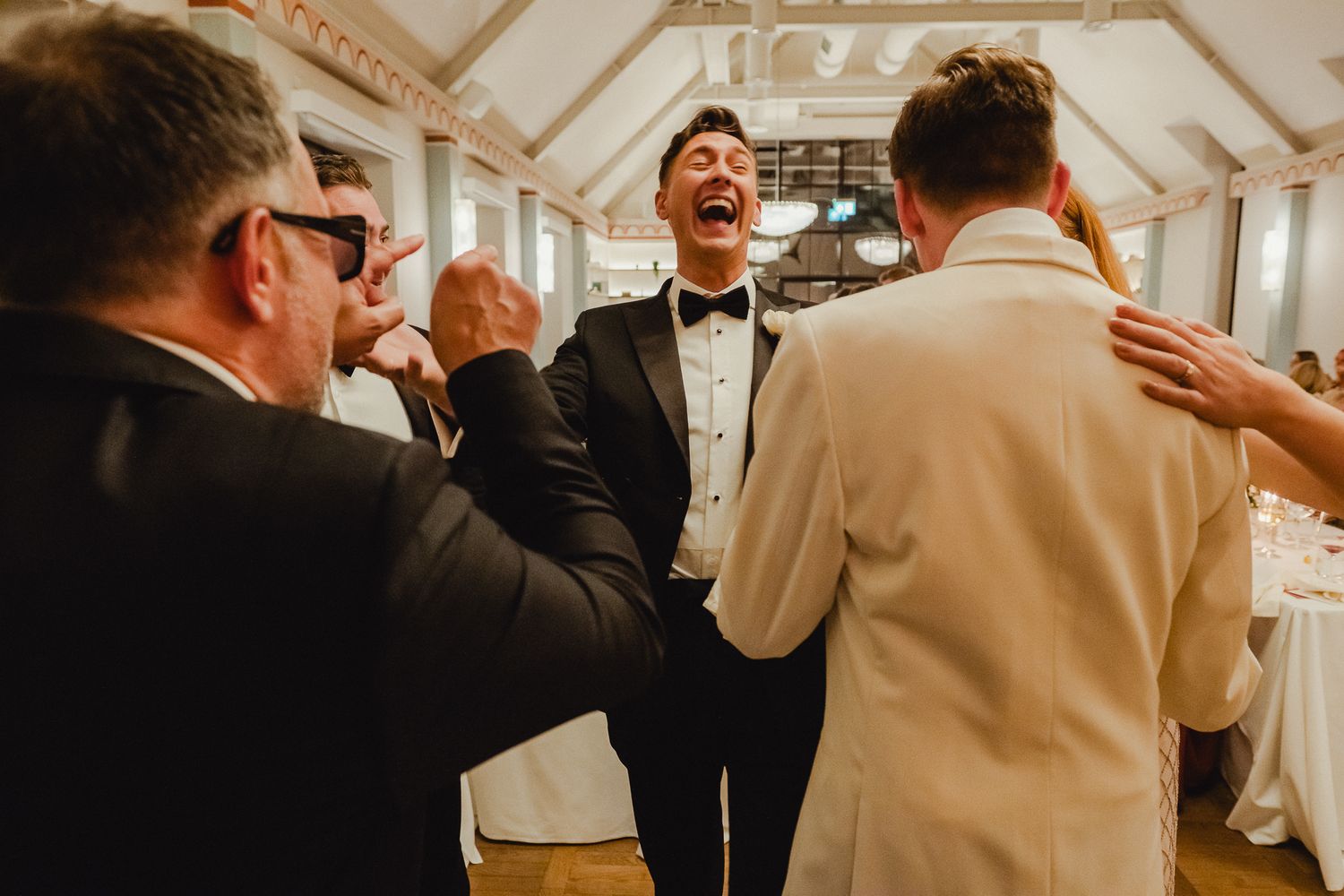 Wedding guests laughing and celebrating during a joyful reception in an elegant white-walled venue.
