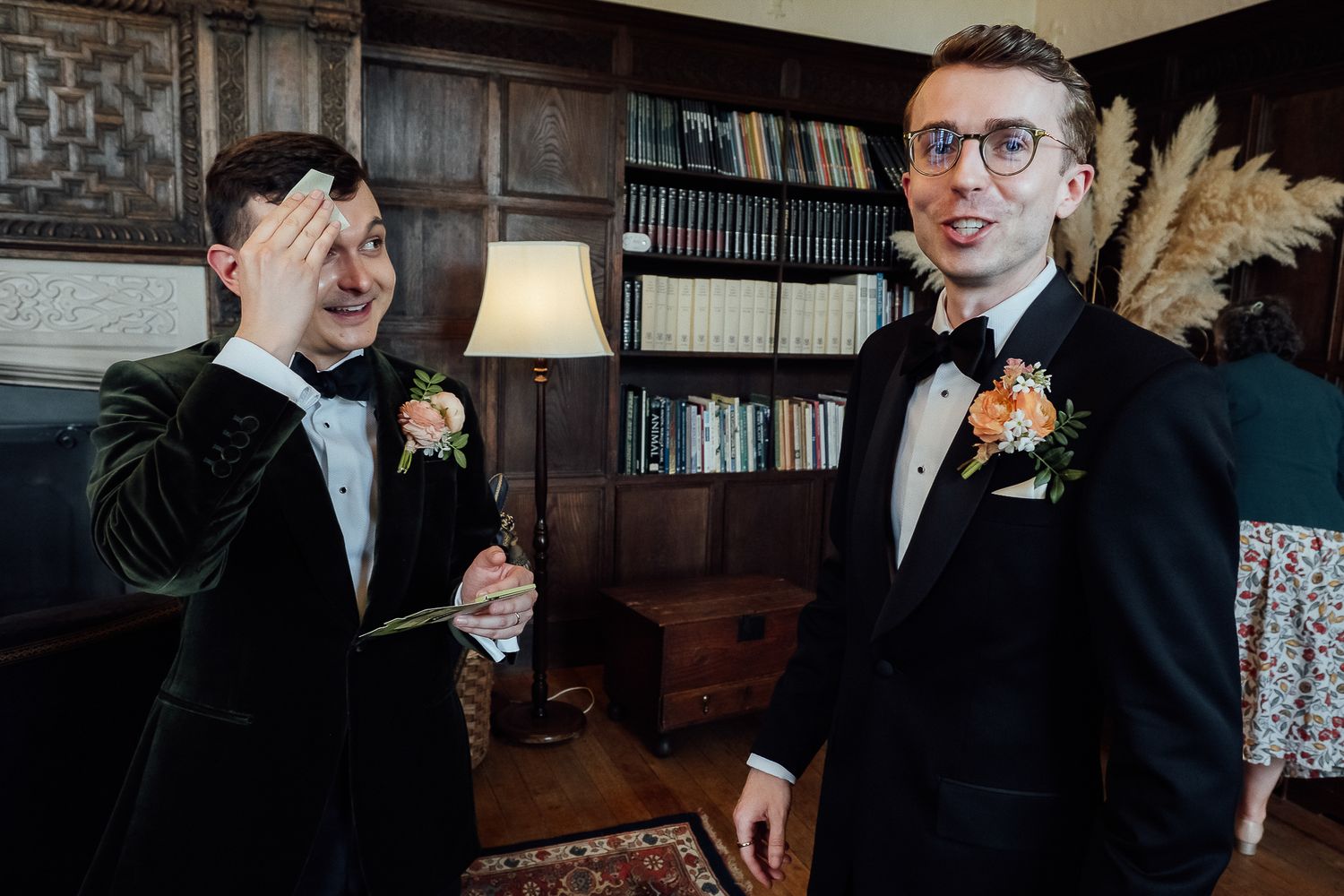 Two grooms in tuxedos with floral boutonnieres share a joyful moment in an elegant wood-paneled library.