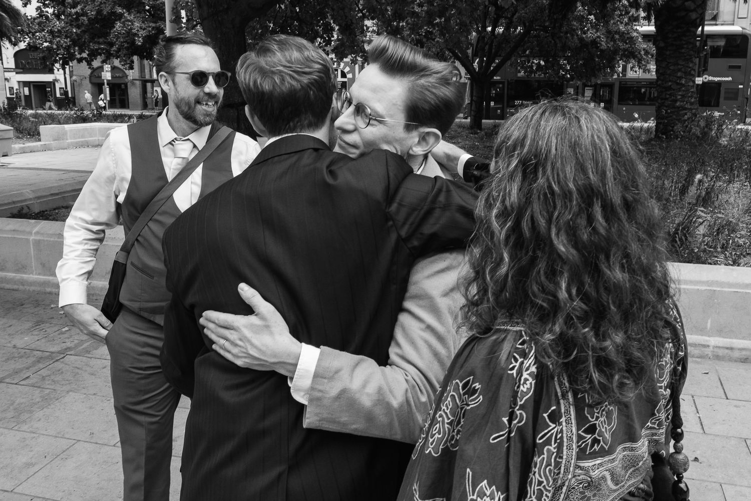Two men in suits share a warm embrace outdoors while a bearded man in sunglasses and a woman with curly hair look on in black and white.