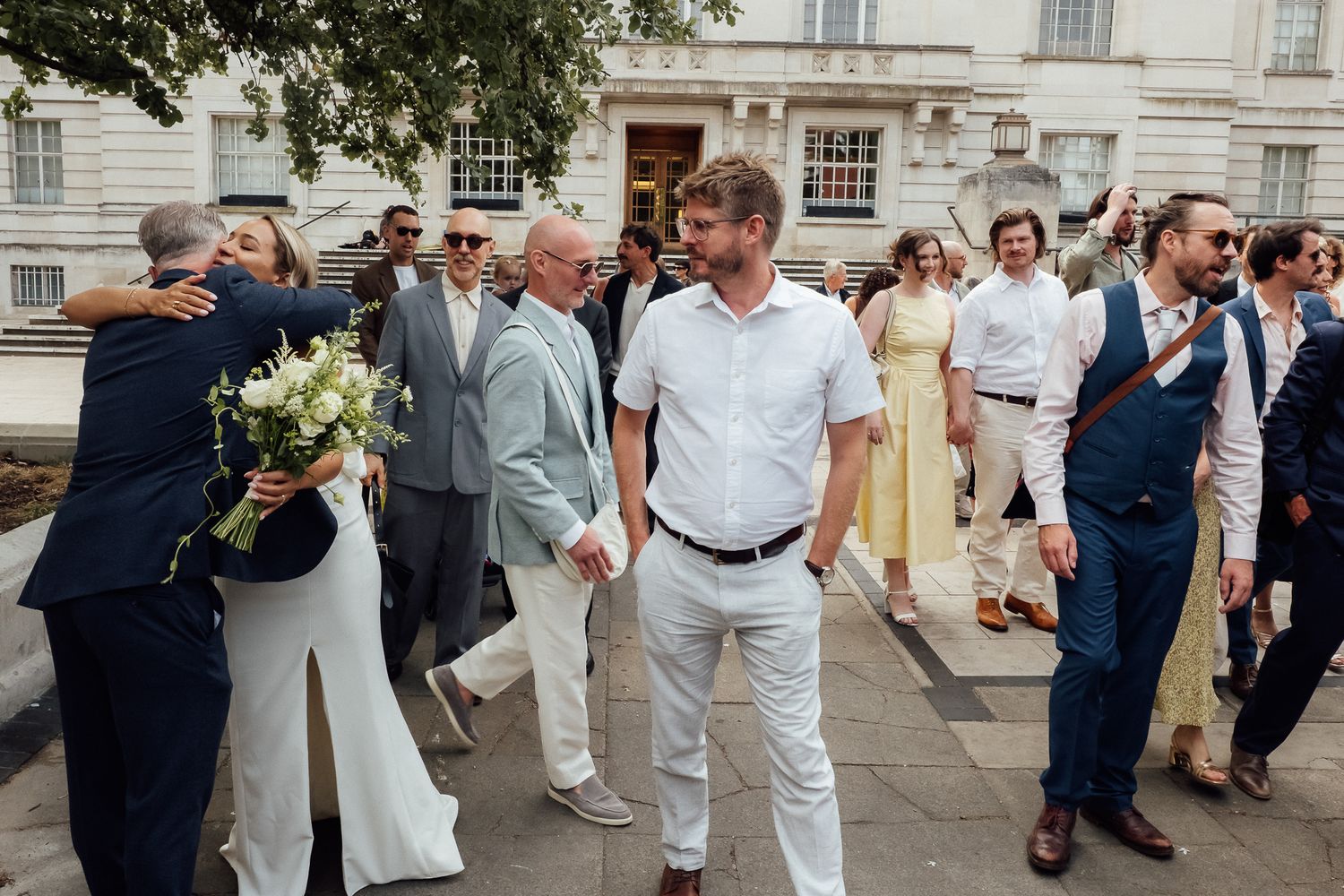 Wedding guests in summer attire gather outside a historic building, with a bride holding a bouquet embracing a man while others smile.