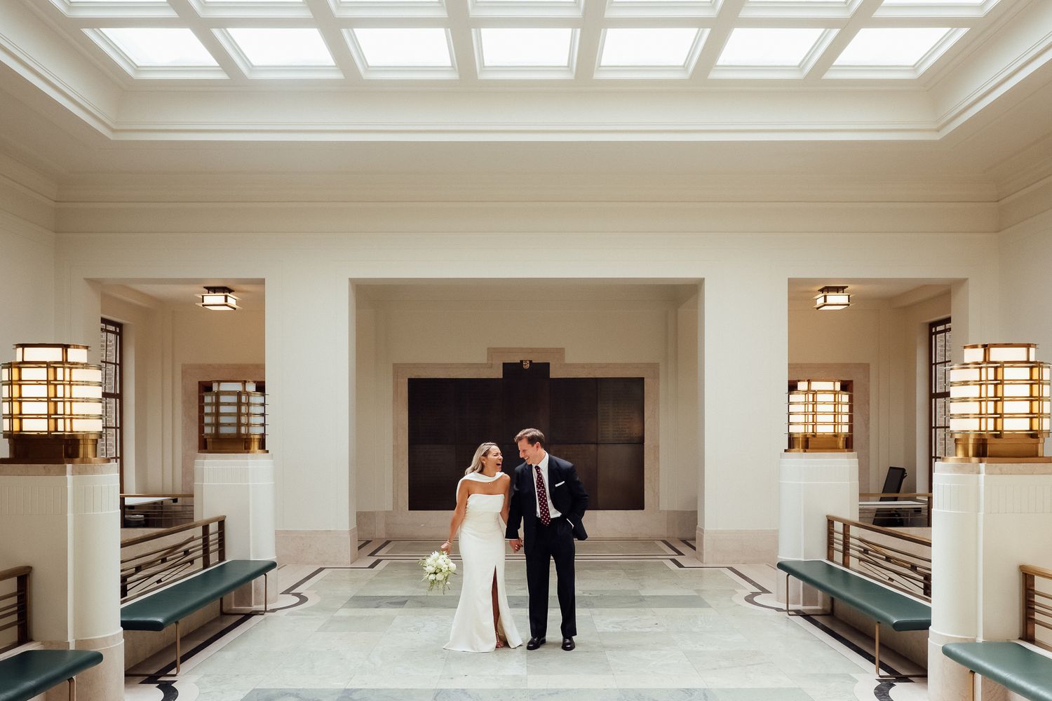 Bride in a white gown and groom in a dark suit embrace in a grand, sunlit hall with skylights, marble floors and teal benches.