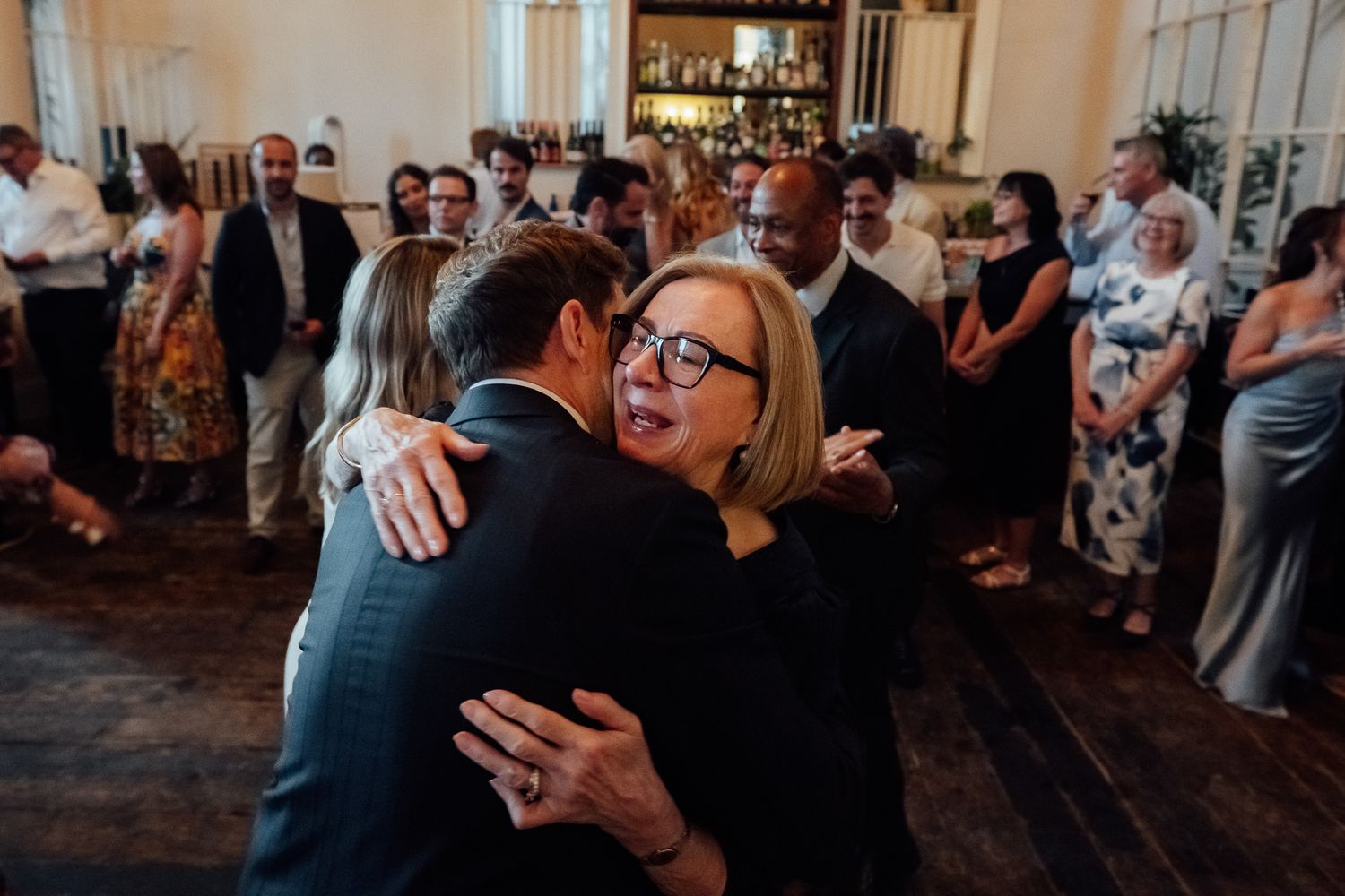A smiling woman with glasses embraces a man in a dark suit while guests mingle in a warmly lit event space behind them.