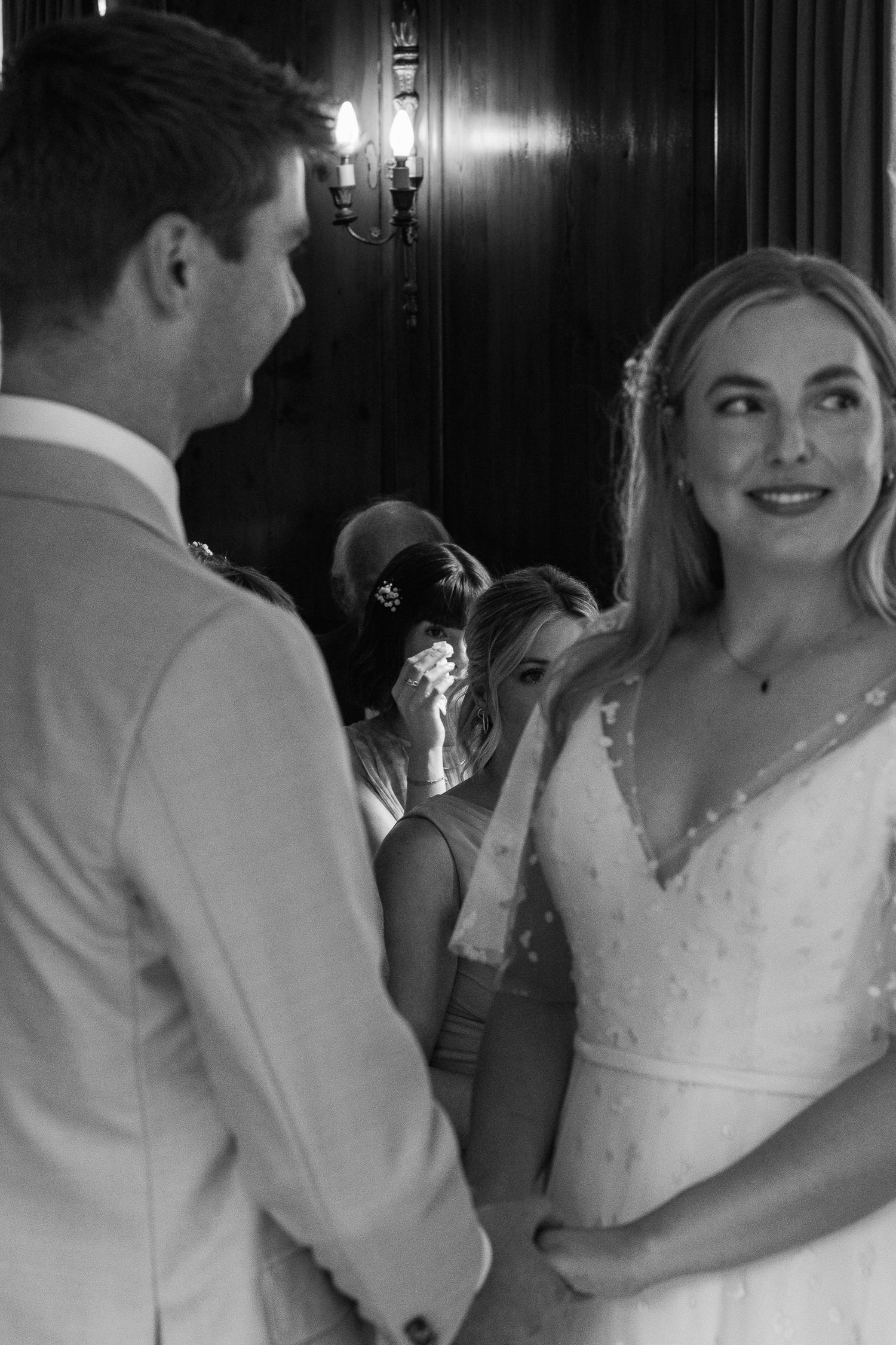 Smiling bride in a lace wedding dress holds hands with the groom during a ceremony as emotional guests look on in black and white.