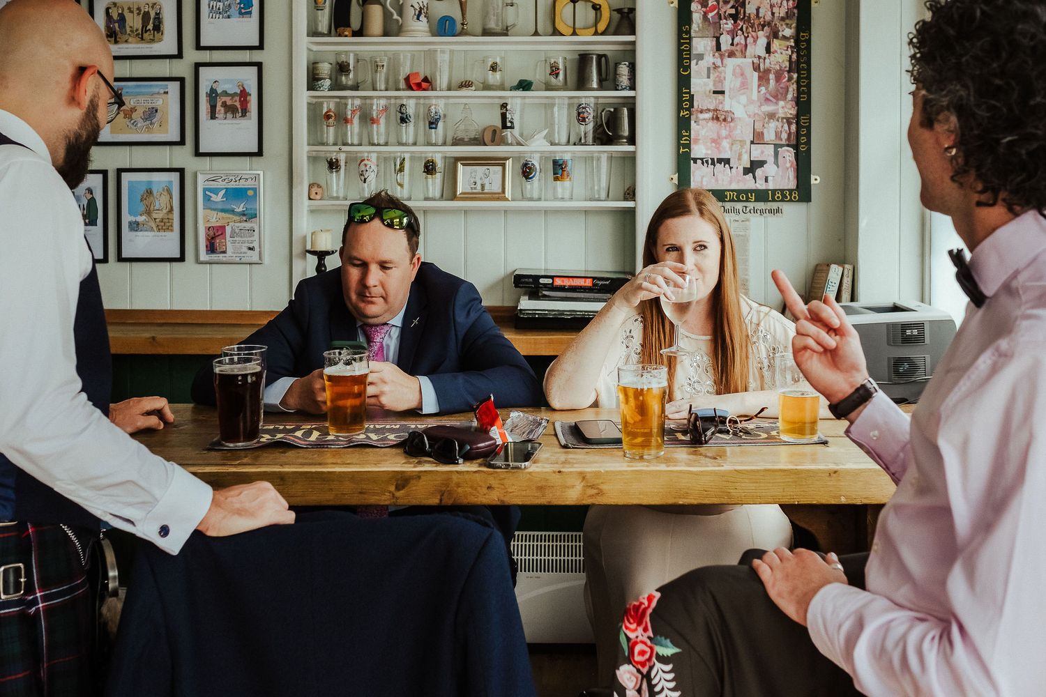 Friends laughing and chatting around a wooden table with pints of beer in a cozy pub with shelves and framed pictures on the walls.