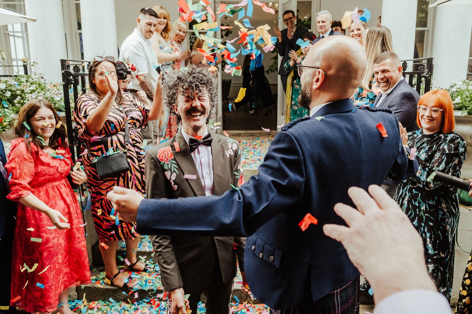 Joyful wedding guests celebrate with colorful confetti as a smiling man in a bow tie and navy suit greets the crowd outdoors.