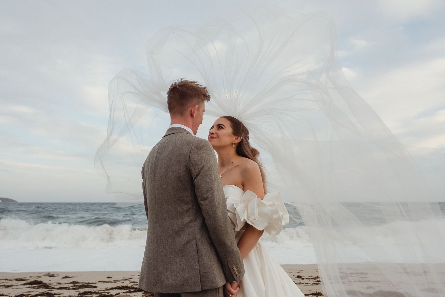 A couple shares a romantic moment on a beach with a flowing white veil caught in the ocean breeze.
