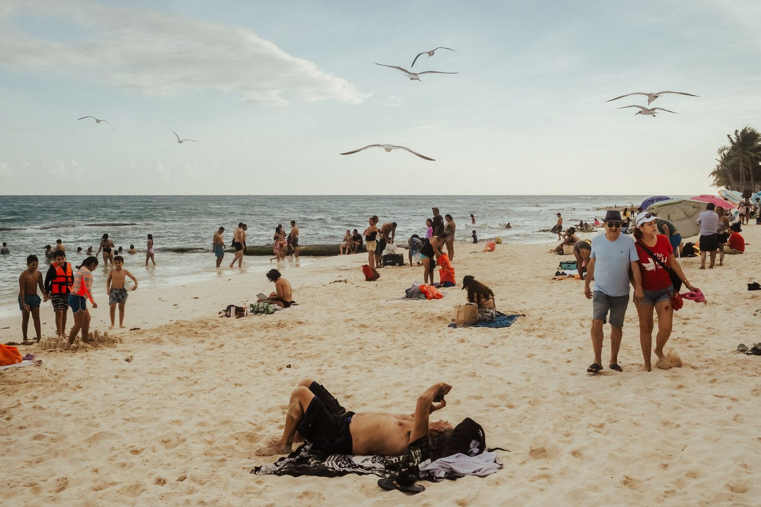 Beachgoers relax and enjoy a sunny day on a sandy beach with seagulls flying overhead near the ocean.