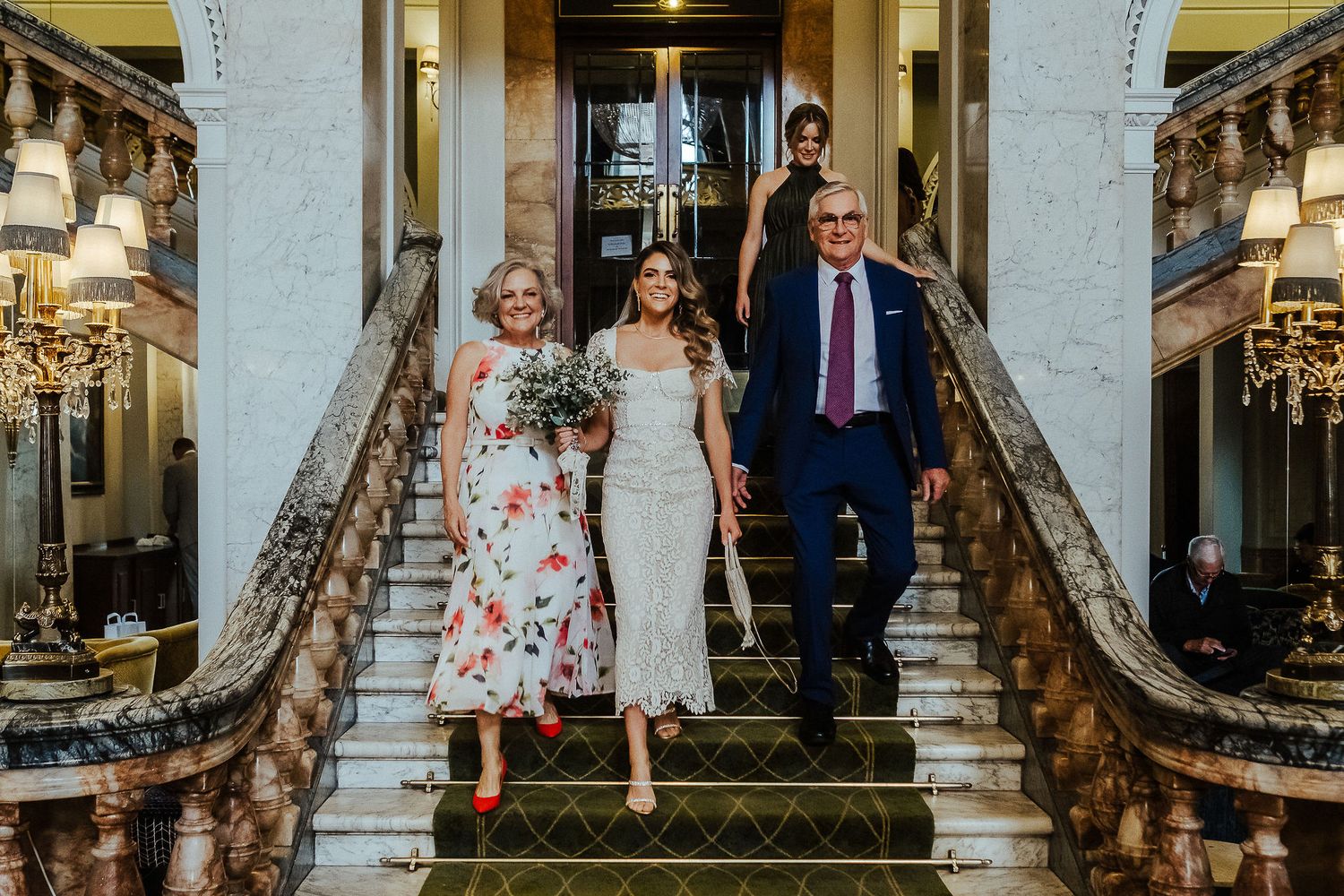 Three people descending an elegant marble staircase with ornate railings in a vintage-style interior.