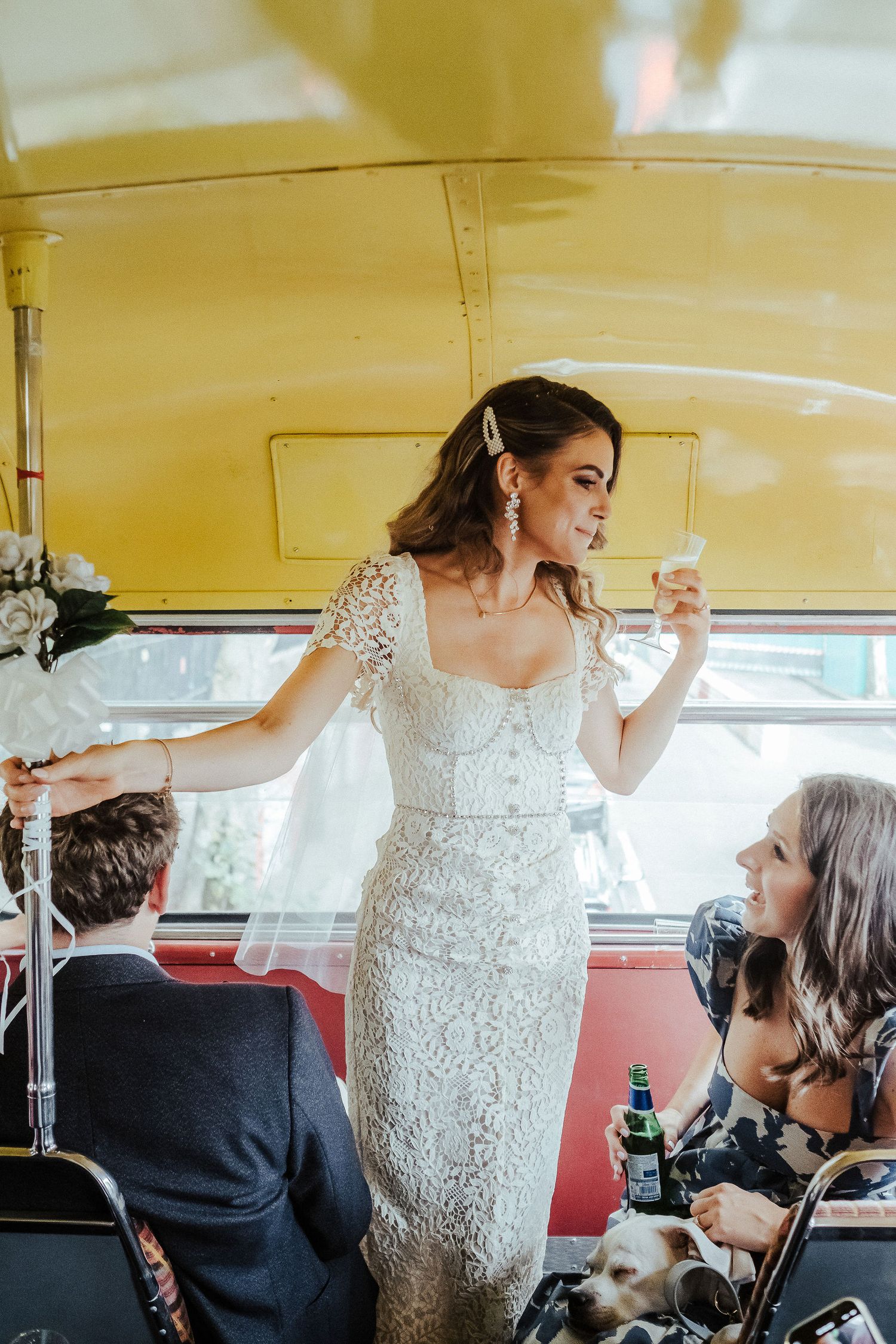 Bride in lace dress celebrates with guests inside a vintage yellow bus during wedding festivities.