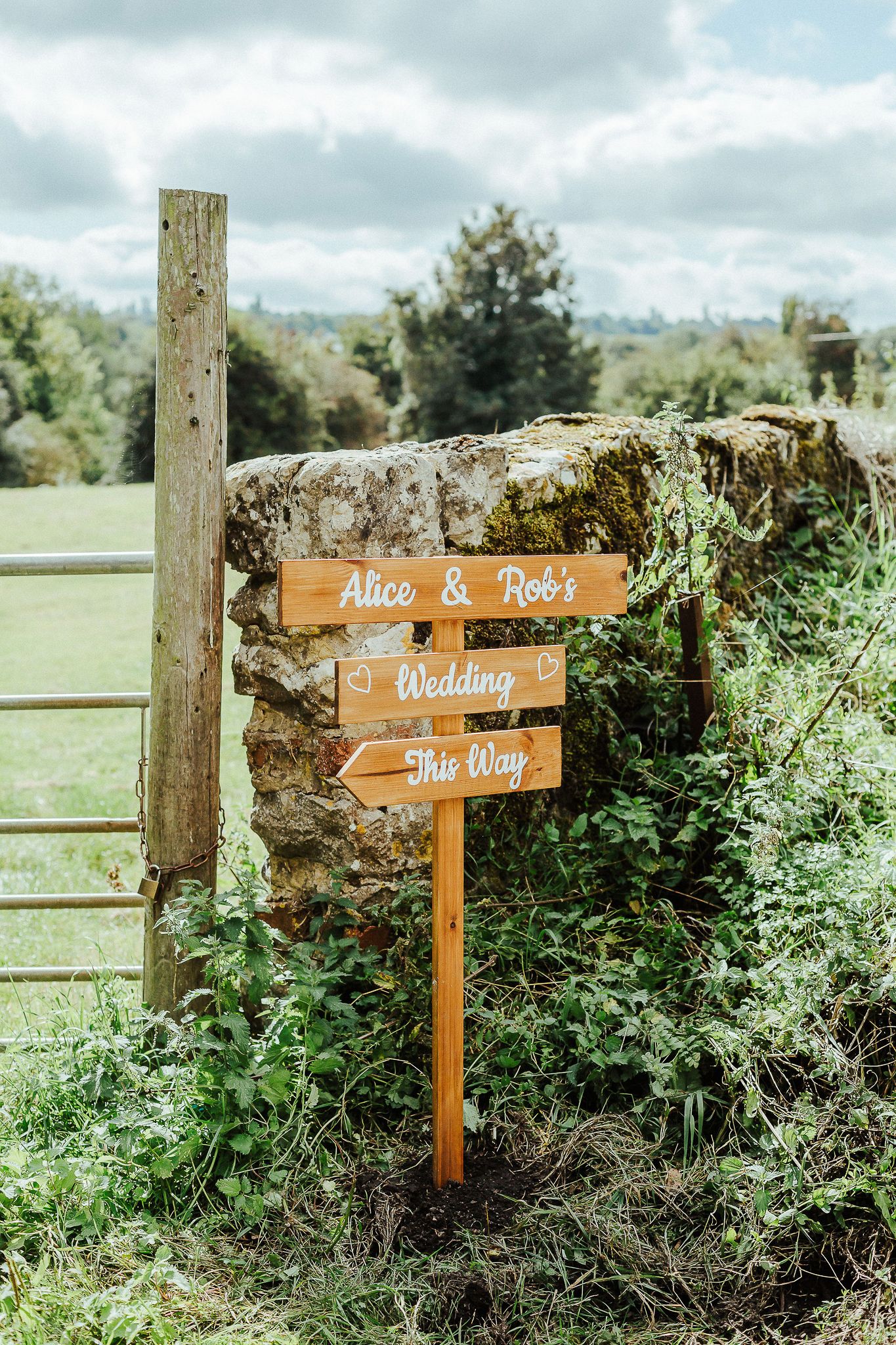 A rustic wooden wedding direction sign mounted on a stone wall in a countryside setting.