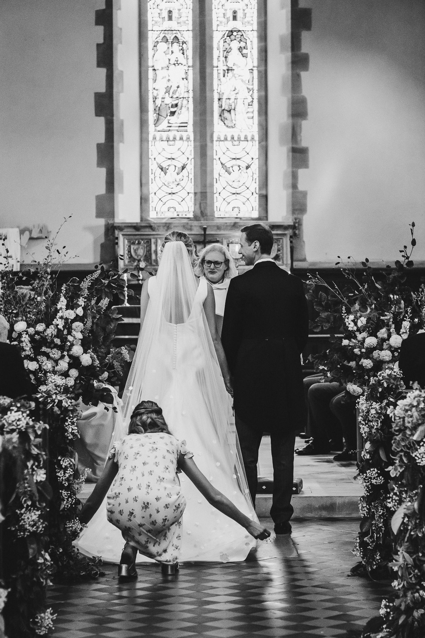 Black and white photo of wedding ceremony in church with stained glass windows and floral decorations.