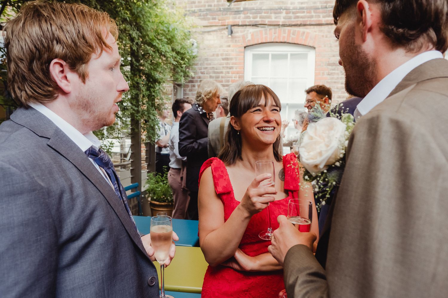 Wedding guests mingle and socialize at an outdoor garden reception while holding drinks and enjoying conversation.