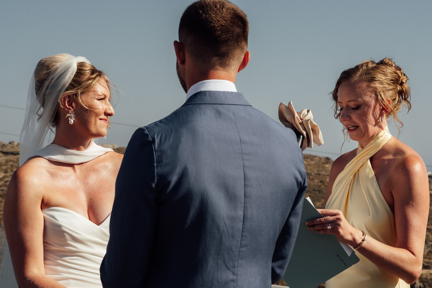 Bride and bridesmaid exchange vows outdoors at a sunny wedding ceremony, with groom in navy suit.