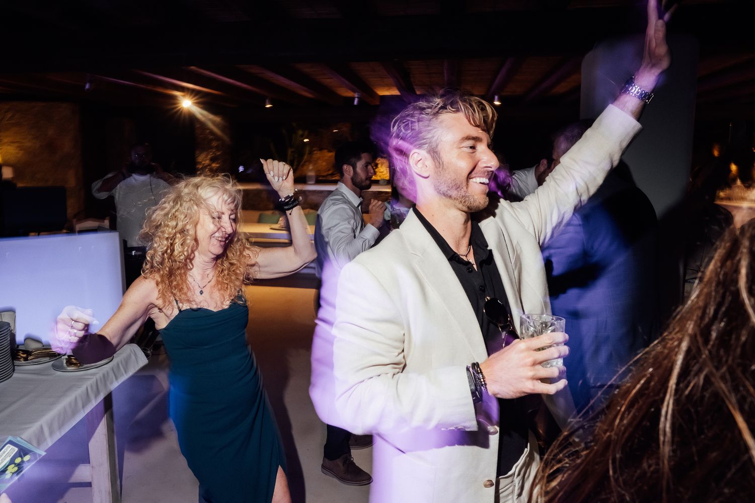 People dancing joyfully at an indoor party, man in white blazer raises arm, woman in teal dress beside him.