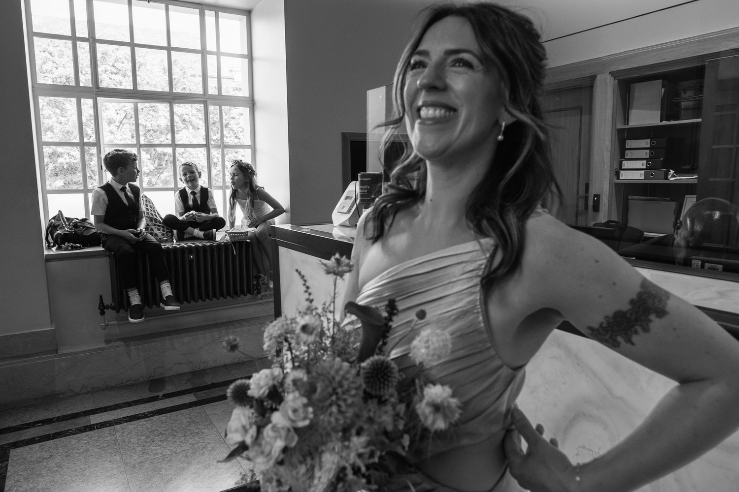 Smiling bride in a strapless gown holds a wildflower bouquet while guests sit on a radiator beside a large window in black and white.