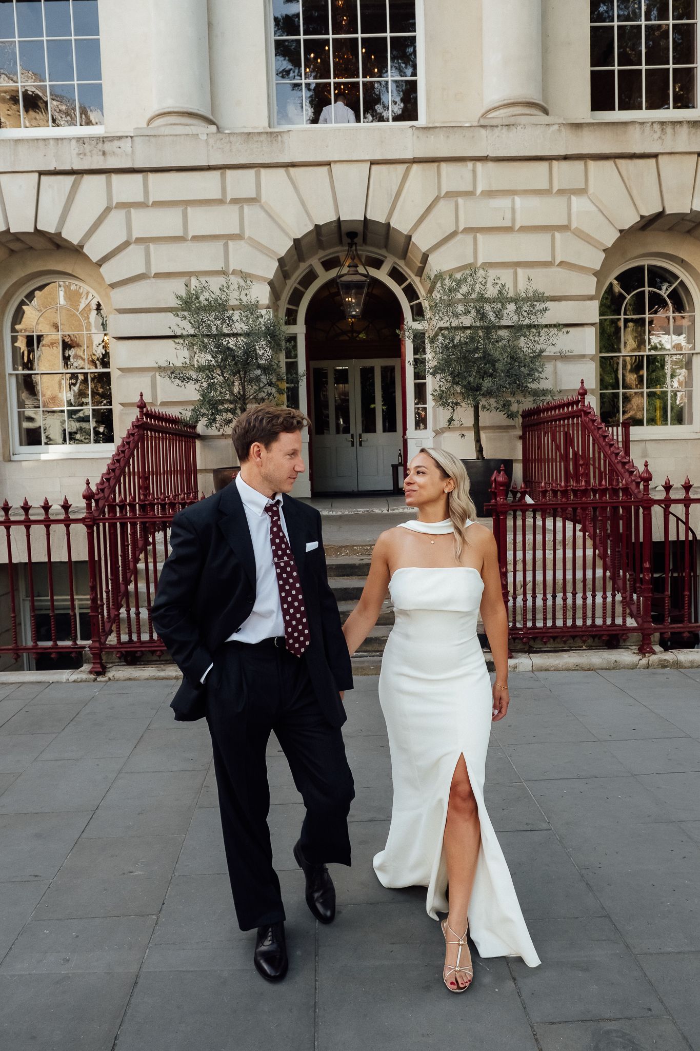 Bride in a white strapless gown with high slit walks hand in hand with a groom in a dark suit and burgundy tie outside a grand building.