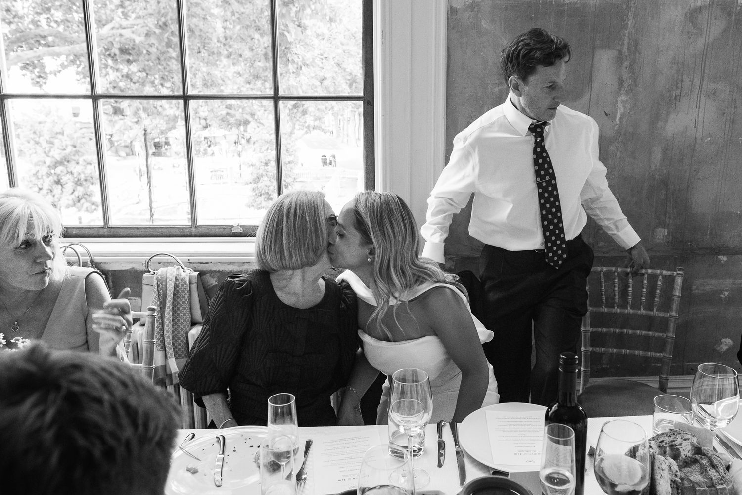 A woman in a dark top kisses another woman on the cheek at a wedding reception table while a man in a tie stands nearby.