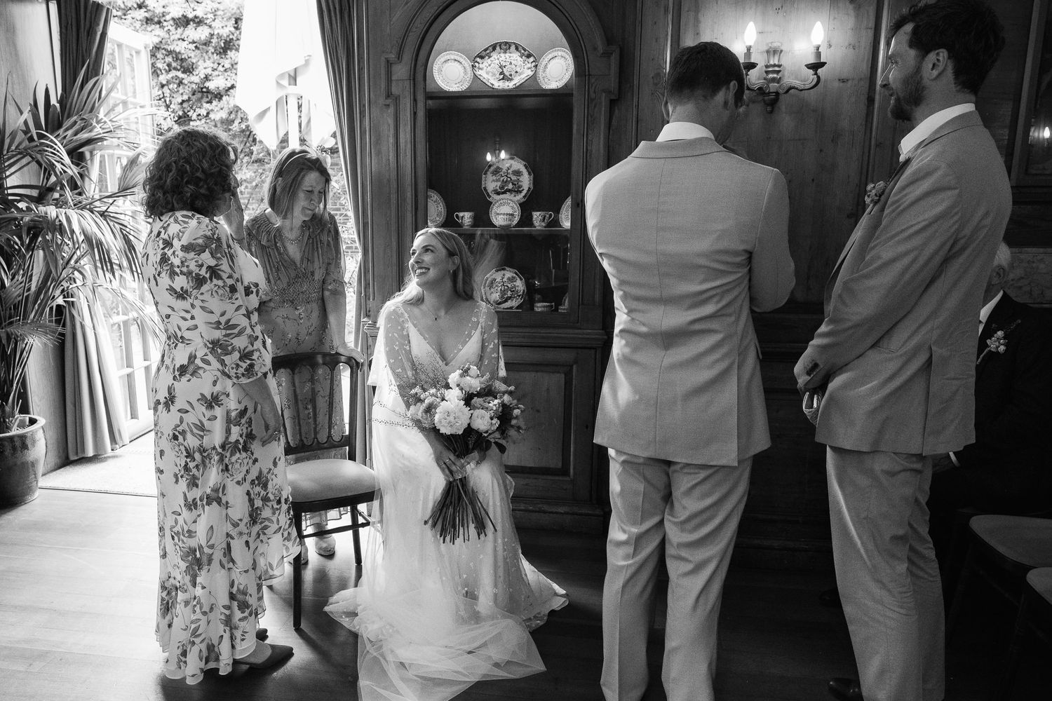 Black and white wedding photo of a bride seated holding a bouquet while two groomsmen stand in front and guests look on.