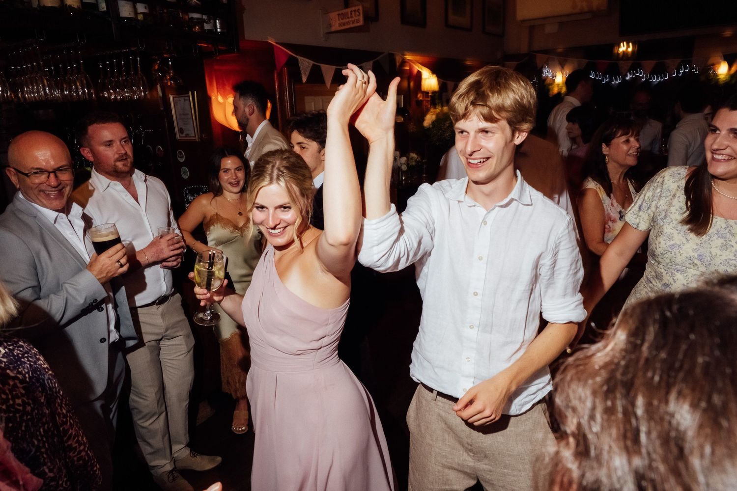 Smiling couple dancing with raised hands at a lively indoor wedding reception while guests cheer and mingle in the warmly lit background.