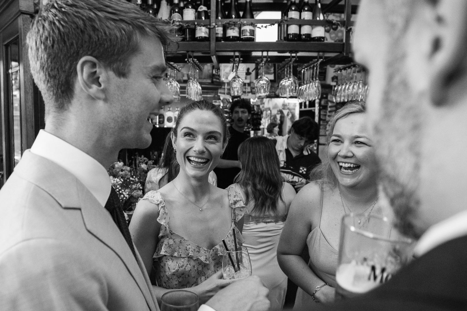 Black and white photo of smiling friends laughing together at a crowded bar with shelves of bottles in the background.