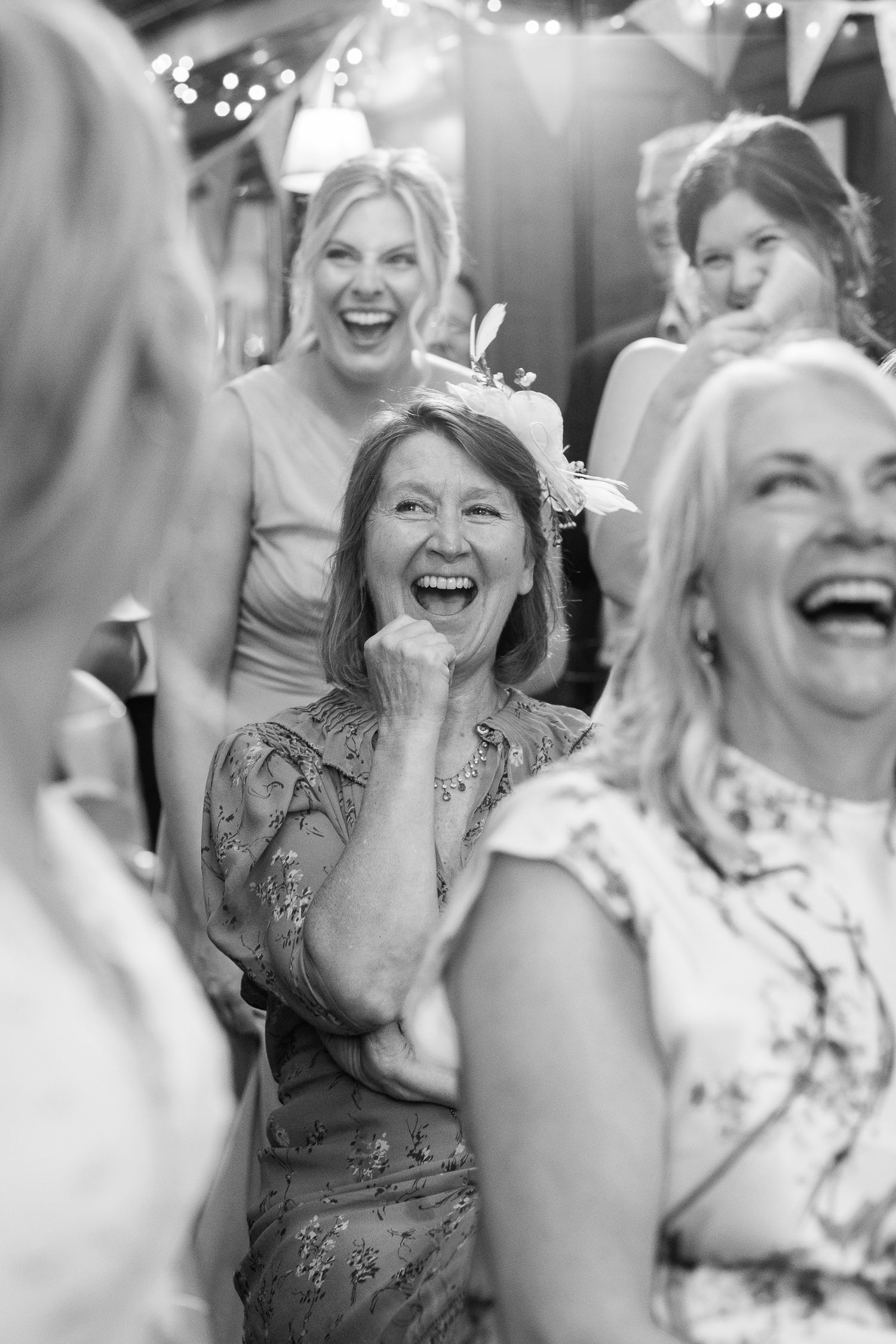 Black and white photo of women laughing joyfully at a celebration, one wearing a floral fascinator, with string lights glowing behind them.