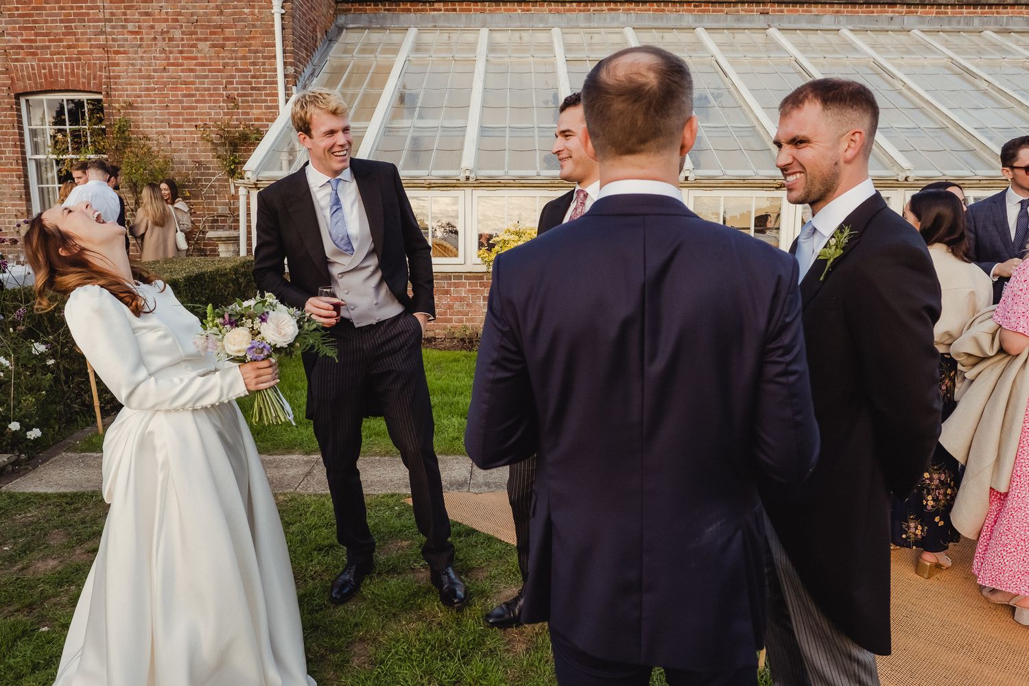 Wedding guests share a lighthearted moment during an outdoor garden reception at a brick venue.
