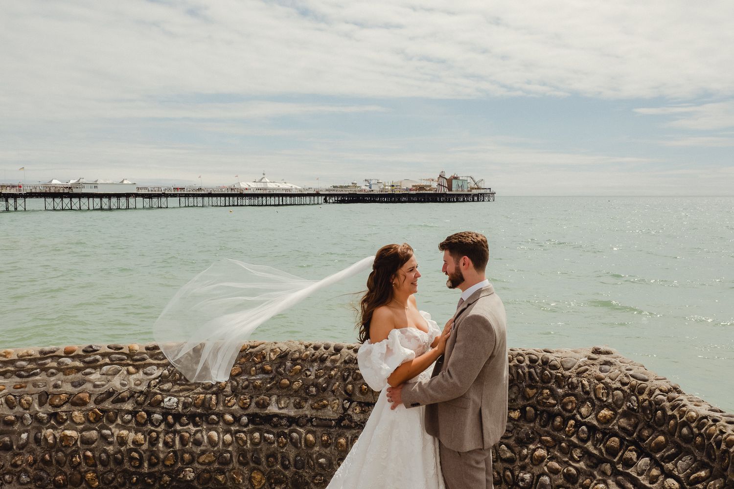 A couple in wedding attire embrace on a rocky beach with a long pier stretching into the turquoise ocean behind them.