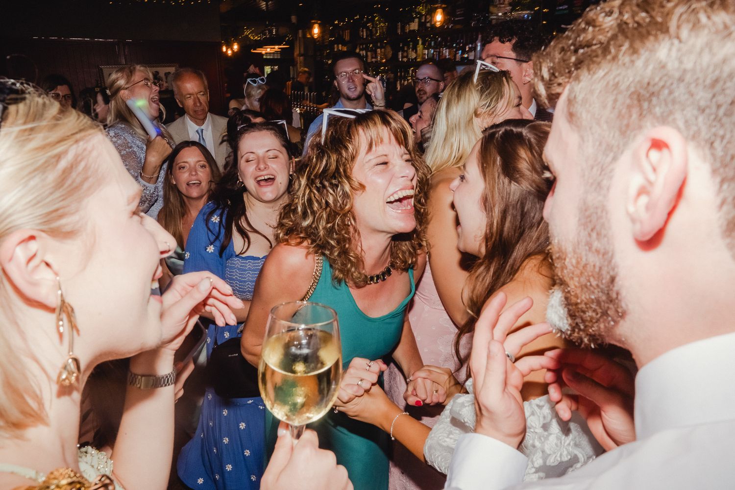 Lively wedding party scene with guests dancing and laughing together at an indoor reception venue.